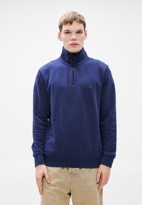 Φούτερ σε ναυτικό μπλε με φερμουάρ μέχρι το στήθος, κατασκευασμένο από απαλό ύφασμα, με ψηλό γιακά, ριμπ μανσέτες και μικρό λογότυπο στο στήθος.