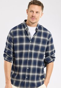 Homme portant une chemise à carreaux bleus et blancs boutonnée sur un t-shirt blanc, manches retroussées, regardant légèrement sur sa gauche.