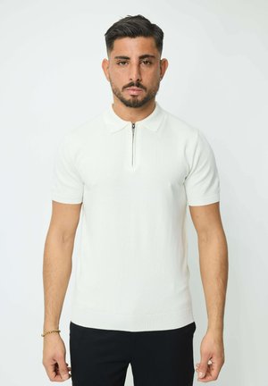 Homme aux cheveux courts et foncés avec une barbe, portant un polo blanc à manches courtes zippé et un pantalon noir, debout devant un fond uni.