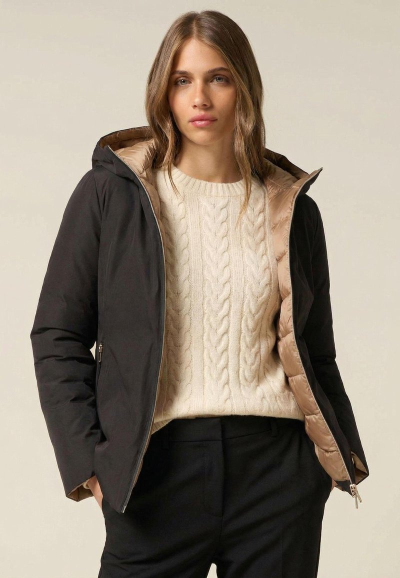 Czarna kurtka puchowa z beżowym wnętrzem, zapinana na zamek, z kapturem, noszona na kremowym swetrze w warkocze. Miękka tekstura i dopasowany fason.