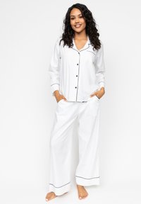 Witte pyjama set met een knoopsluiting bovenaan voorzien van zwarte piping en lange mouwen, gecombineerd met losse, wijde broekspijpen die ook zijn afgewerkt met zwarte randen.
