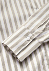 Colletto della camicia a righe con strisce marroni, gialle e bianche; tessuto di cotone; singolo bottone bianco sul polsino; superficie liscia.