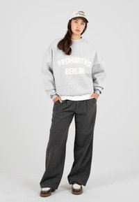 Fiatal nő szürke pulóverben, amelyen a "PROHIBITED BERLIN" felirat szerepel, sötét, bő szárú nadrágban, fehér loaferekben és bézs baseball sapkában áll, kezét a zsebébe dugva.