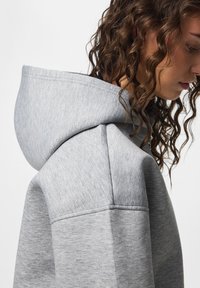 Giovane donna con capelli castani ricci che indossa una felpa con cappuccio grigio chiaro, vista di profilo su uno sfondo semplice.