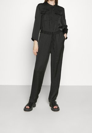 Vrouw draagt een zwart jumpsuit met lange mouwen en een taille met trekkoord, en zwarte chunky sandalen met open teen, staand tegen een witte achtergrond.