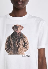 Bílý bavlněný tričko s potiskem hnědého psa v hnědé bundě. Pod obrázkem je text "BARBOUR".