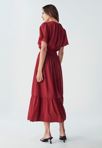 Crvena polusvijetla maxi haljina s kratkim lepršavim rukavima, naboranim strukom i volanima na rubu. Uparena s crnim sandalama na visoku petu.