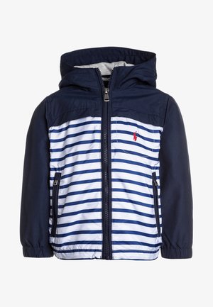 Marineblå og hvidstribet jakke med hætte, der har lynlås foran og to sidelommer. Materialet ser let og glat ud. Rød logodetalje.