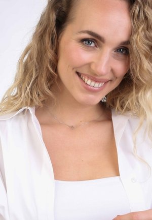 Silberne Halskette mit einem herzförmigen Anhänger, getragen über einem weißen Oberteil. Das Model hat welliges Haar und lächelt. Der Hintergrund ist schlicht weiß.