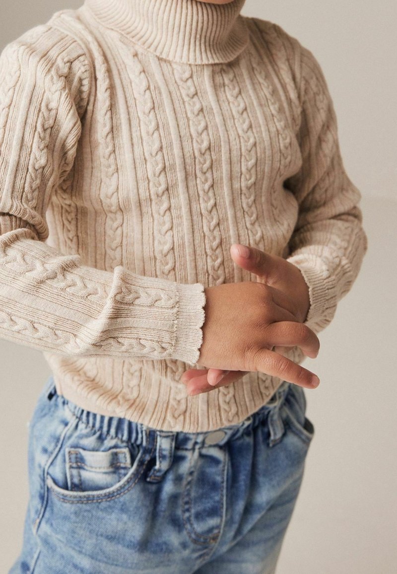 Enfant portant un pull à col roulé en maille torsadée beige et un jean bleu, les mains croisées légèrement à la taille.