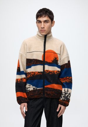 Jeune homme portant une veste polaire beige avec une fermeture éclair noire, présentant un motif de paysage coloré avec un soleil orange et des rayures bleues, noires et marron.