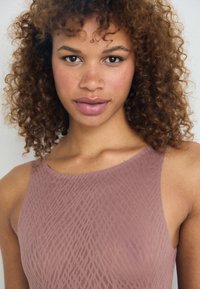 Mauve sleeveless top with a textured, wavy pattern. Model has curly hair and natural makeup, focusing on facial features and expression.
