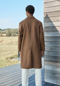 Brauner Wollmantel mit maßgeschneiderter Silhouette, langen Ärmeln, Reverskragen und Schlitzen, getragen über weißen Hosen.