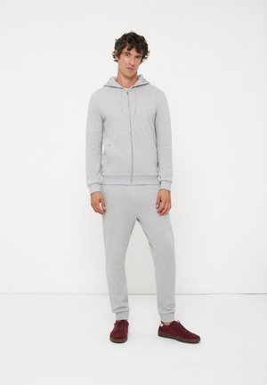 Homme debout portant un sweat à capuche zippé gris clair et un jogging avec des baskets rouge foncé sur fond blanc uni.