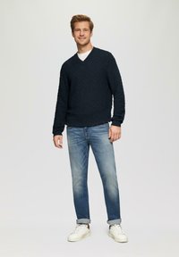 Mann in einem strukturierten marineblauen Pullover, weißem T-Shirt, ausgewaschenen Jeans mit Umschlägen und weißen Sneakers, der vor einem schlichten hellen Hintergrund steht.