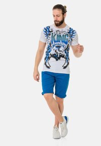 Weißes T-Shirt mit kurzen Ärmeln und blauem Grafikdesign, das "KING" sowie Designs in Schwarz und Blau zeigt. Kombiniert mit blauen, hochgekrempelten Shorts und weißen Sneakers.