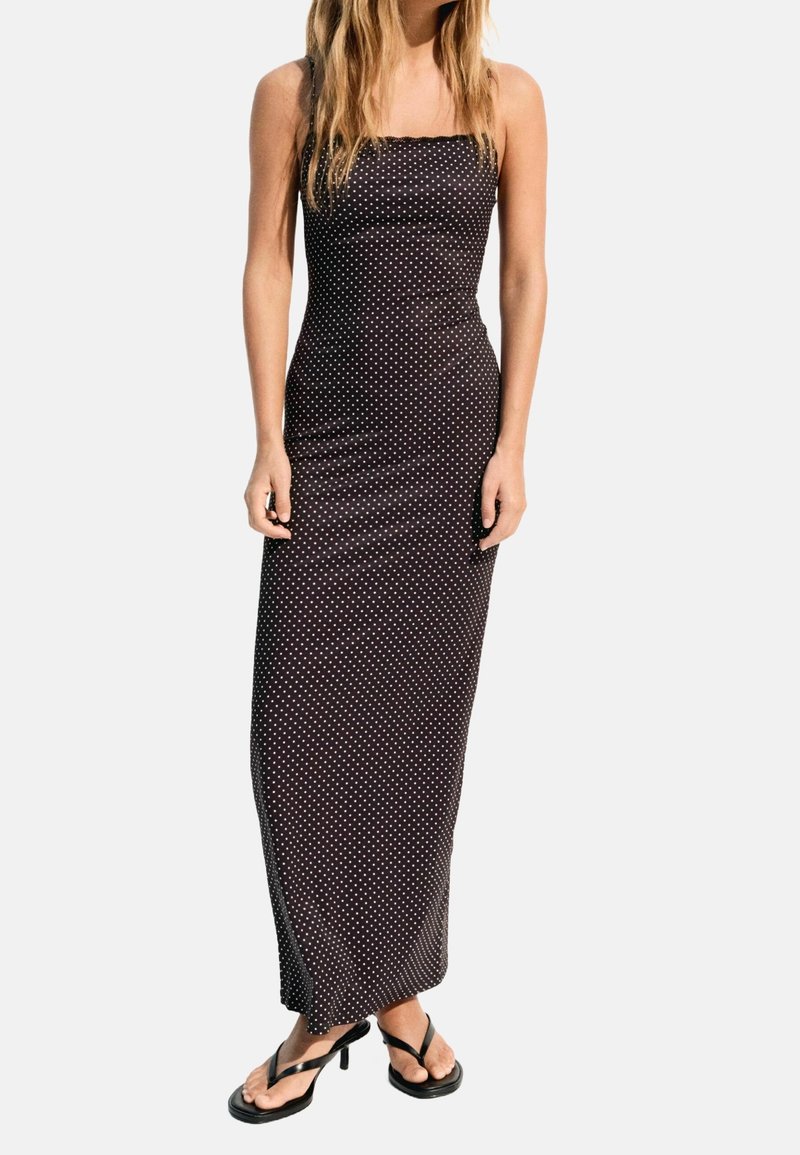Femme portant une robe maxi noire sans manches à pois blancs et des sandales noires à talons, debout devant un fond uni.