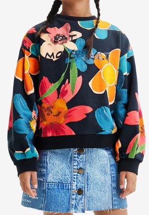 Enfant portant un sweat noir avec de grandes fleurs colorées et du texte, associé à une jupe en jean bleue boutonnée avec des détails cousus.
