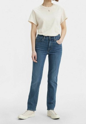 Person wearing a plain white short-sleeve shirt, high-waisted blue jeans, white sneakers, with one hand in pocket against a white background.