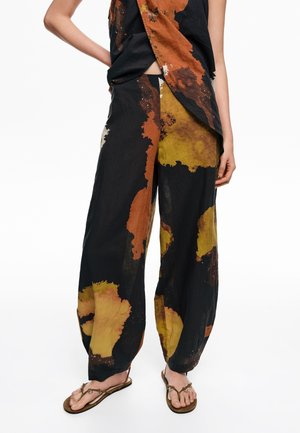 Pantalones negros sueltos con grandes salpicaduras abstractas en naranja y amarillo, combinados con un top asimétrico a juego y sandalias marrones en una persona de pie.
