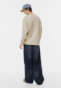Cămașă bej cu mânecă lungă, cu un croi lejer, asortată cu blugi evazați de culoare albastru închis. Modelul poartă o șapcă bicoloră cu visieră.