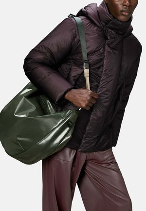 Giubbotto imbottito in tessuto viola scuro con colletto alto e cuciture visibili; tiene in mano una borsa lucida verde scuro con tracolla.