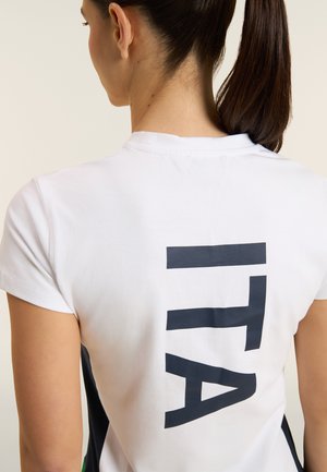 Donna con coda di cavallo scura che indossa una camicia bianca con grandi lettere blu navy "LTA" stampate verticalmente sulla schiena.