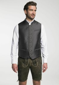 Schwarze gemusterte Weste mit hohem Kragen, Knopfleiste und zwei Taschen, getragen über einem weißen Hemd und kombiniert mit grünen Shorts.