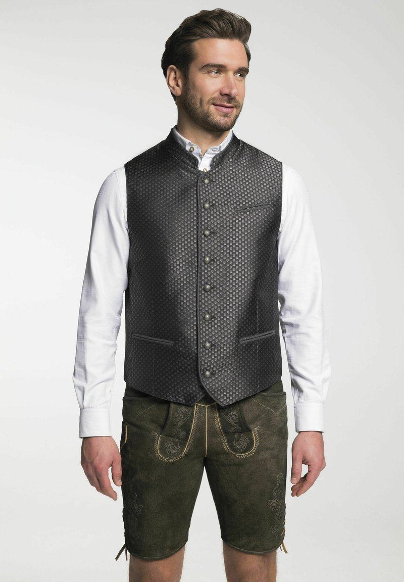 Schwarze gemusterte Weste mit hohem Kragen, Knopfleiste und zwei Taschen, getragen über einem weißen Hemd und kombiniert mit grünen Shorts.