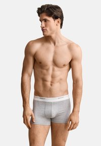 Jeune homme aux cheveux courts et foncés, portant un boxer gris GANT, debout torse nu et regardant vers sa gauche sur un fond clair uni.