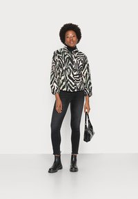 Casaco com padrão de zebra em preto, branco e verde, combinado com jeans skinny pretos e botas de tornozelo pretas, segurando uma bolsa preta.