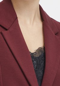 Blazer bordeaux avec un col cranté. En dessous, un haut en dentelle noire est visible, mettant en valeur des motifs floraux complexes et des textures délicates.