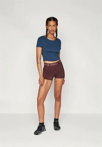 Ženska v športni obleki s spletenimi lasmi, oblečena v modro kratko majico, rjave Nike Pro kratke hlače, črne nogavice in črne superge.