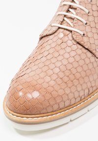 Gros plan d'une chaussure en cuir marron clair avec une texture ressemblant à des écailles, des lacets blancs et une semelle blanche avec des détails de couture au niveau des bords.