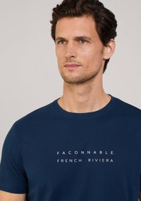 Camiseta de algodón azul marino de manga corta, con el texto impreso en blanco "FAÇONNABLE FRENCH RIVIERA" centrado en la parte delantera. Textura suave.
