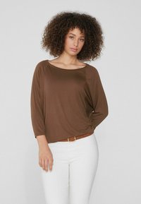 Braunes Langarmoberteil mit weiter Ausschnitt und Raffung an der Seite, kombiniert mit weißen Hosen und einem braunen Gürtel. Sanfte Stofftextur.