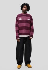 Maroon striped long-sleeve shirt with white graphics, paired with black oversized jeans and yellow boots. Casual, relaxed fit.