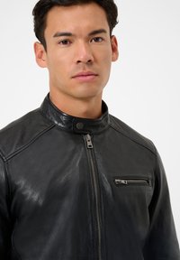 Schwarze Lederjacke mit Stehkragen, Frontreißverschluss und einer Reißverschlusstasche auf der Brust. Weiche Textur und glatte Oberfläche, taillierte Passform.