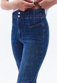 Högmidjade denimjeans med en mörkblå tvätt, dubbla framfickor och två guldfärgad knappar i midjan. Slät textur.