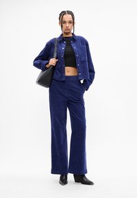 Γυναίκα με πλεγμένα μαλλιά φοράει μια μπλε βελουτέ ζακέτα και παντελόνι, μαύρο κοντό τοπ, μαύρες ψηλές μπότες και κρατά μια μαύρη τσάντα ώμου.