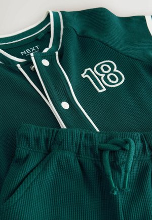 Grüner Sweatshirt mit Struktur, weißer Einfassung und Druckknöpfen, mit einer bestickten "18"; kombiniert mit grünen Shorts mit elastischem Bund.