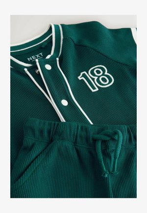 Zaļš teksturēts džemperis ar baltu apmali un sprādzēm, uzšūts ar izšuvumu "18"; komplektā ar zaļiem šortiem ar elastīgu jostasvietu.