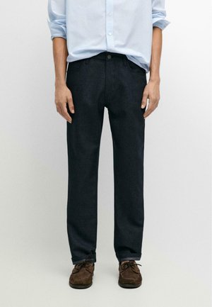 Person iført mørke jeans med lige ben, lyseblå stribet skjorte med opkørte ærmer og brune mokkasinsko, stående mod en hvid baggrund.