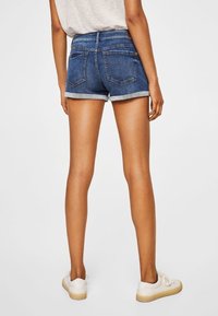 Denim shorts met een donkerblauwe wassing, opgerolde zoom en twee achterzakken. Gestyled met een lichtgrijze top en witte sneakers.