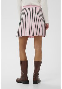 O femeie îmbrăcată cu o fustă mini roz cu dungi verticale negre și cizme din piele maro, până la jumătatea gambei, stând pe un fundal alb.