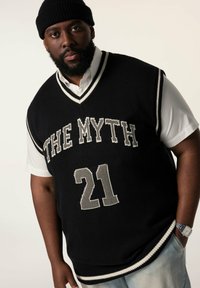 Schwarze Strickweste mit weißem Saum, mit dem Text "THE MYTH" und "21" in Grau. Tragen über einem weißen Hemd mit Kragen, kombiniert mit hellen Jeans.