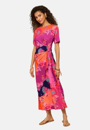 Vrouw met krullend haar, gekleed in een levendige midi-jurk met bloemen in roze, oranje en marineblauw, met korte mouwen en gouden sandalen.