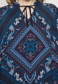 Marinblå topp med paisleymönster i blått, rött och vitt, med v-ringning och knytning vid halsringningen. Mjuk, slät tyg.
