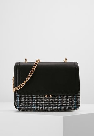 Schwarze Umhängetasche aus Leder und kariertem Stoff mit goldener Kettenriemen, präsentiert auf weißem Podest vor grauem Hintergrund.