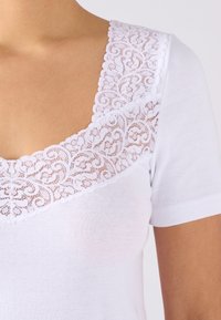 Witte t-shirt met korte mouwen, featuring een gekartelde kantenkraag met een bloemmotief. De stof is glad, wat zorgt voor een zachte textuur.